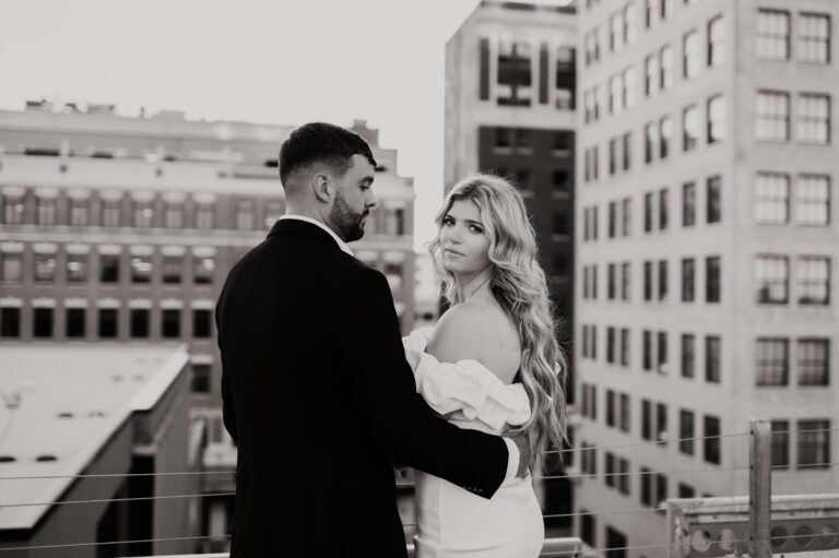 couple on top of rooftop bride looking back at camera while groom looks out into the city skyline black and white image