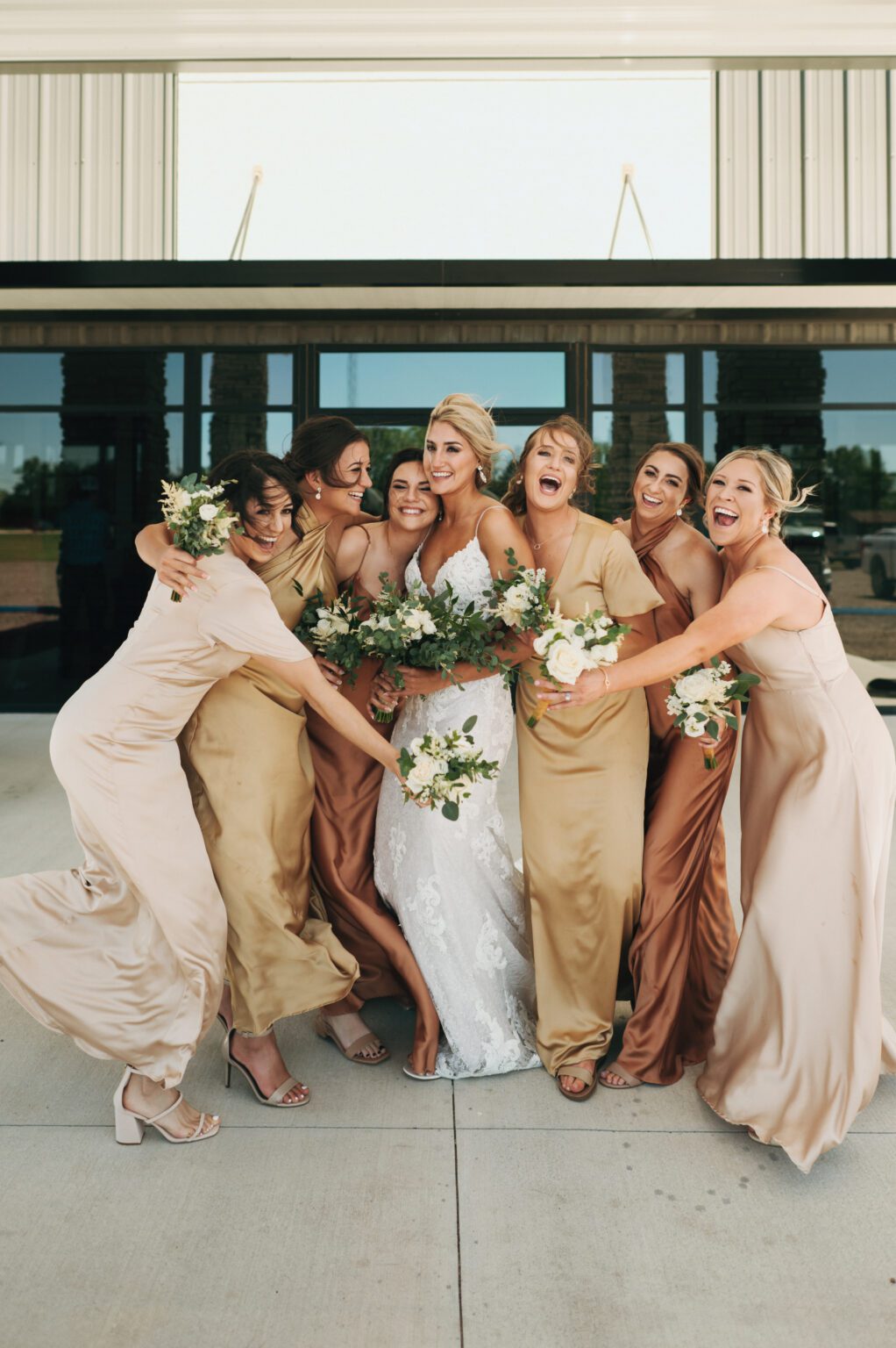bridesmaids hug and laugh with bride in front of church wearing brown and gold silk bridesmaid dresses
